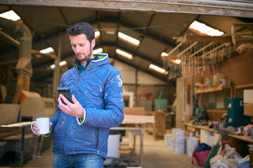 Carpenter Working In Woodwork Workshop Looking At Mobile Phone On Coffee Break