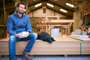 Carpenter Working In Woodwork Workshop With Pet Dog On Coffee Break