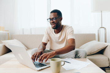 Smiling African American Freelancer Working on Laptop while Sitting on Sofa in Modern Home Office The young black man, dressed in casual attire, is typing on his computer with a focused expression