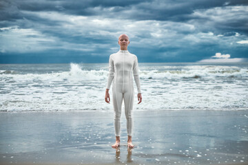 Happy hairless girl with alopecia in white futuristic suit stands on beach bathed by ocean waves,...