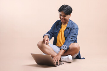 Full Length Of Sitting Young Asian Guy Looking And Pointing At Laptop Isolated On Beige Background