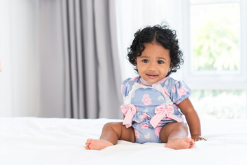 Cute African little newborn 7 months old baby girl wearing clothes with black curly hair sitting on white bed in bedroom at home. Happy adorable small daughter smiling. Copy space