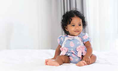 Fototapeta premium Cute African little newborn 7 months old baby girl wearing clothes with black curly hair sitting on white bed in bedroom at home. Happy adorable small daughter smiling. Copy space