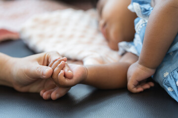 Selective focus on hand of cute African little newborn 7 months old baby girl with black curly hair holding her mother finger while sleeping on sofa with blanket as pillow at home. Family bonding