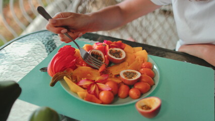 A person is seated at a table, skillfully eating a plate of assorted fruits. The scene shows a variety of fruits being sliced into smaller pieces, exotic fruits