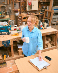 Female Carpenter Working In Woodwork Workshop With Cup Of Coffee Making Notes On Clipboard