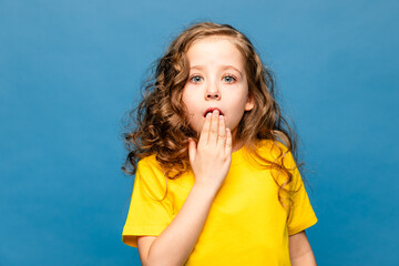 Scared beautiful Caucasian little girl wearing yellow T-shirt over blue background, covering her...