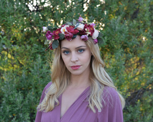 Naklejka premium close up portrait of pretty blonde female model wearing a flower crown wreath and purple dress. green nature plants and trees in background