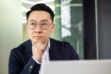 Asian mature man in a suit and glasses thinking and working at an office desk. Business concept.