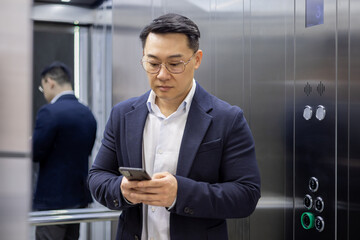 Asian mature man in business attire using a smartphone while standing inside an elevator. Business professional commuting and staying connected.