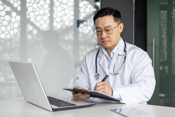 Mature Asian doctor in white coat working on a laptop and writing in a clipboard at the office desk. Professional healthcare and medicine concept.