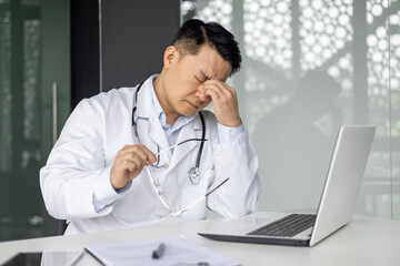 Asian mature doctor looking stressed while sitting at desk with laptop and holding glasses,...