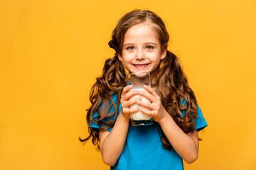 Happy smiling little girl in blue tank top holding a glass of milk on a light yellow background,...
