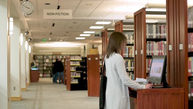 A woman in a light coat searches a library catalog on a computer located in the non-fiction section of a well-stocked library, emphasizing the role of technology in accessing information.