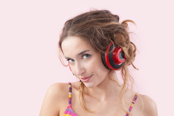 Portrait of young smiling woman who is listening to music through red headphones, looks into the camera with her blue eyes, while being isolated on a pink background
