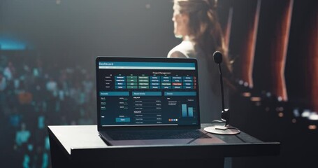 Laptop Computer with a Project Management System Standing on a Podium with a Microphone. Anonymous Blurred Speaker Standing in the Background, Making a Tech Demonstration of the Software