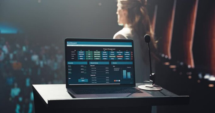Laptop Computer with a Project Management System Standing on a Podium with a Microphone. Anonymous Blurred Speaker Standing in the Background, Making a Tech Demonstration of the Software