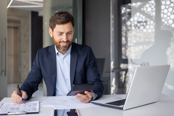 Smiling young male accountant, financial expert and analyst sitting in modern office at desk, working with documents and calculator