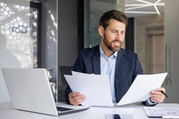 A smiling young man works in the office with documents, sits at a desk with a laptop, checks and...
