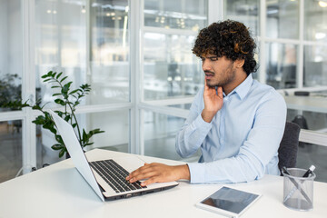 Man suffering from toothache while working on his laptop in a modern office setting. Pain and...