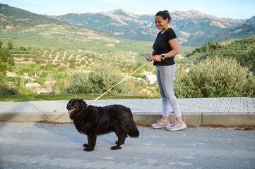 Side portrait of a woman in sportswear, walking her dog on leash in the nature. Multi ethnic...