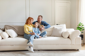 Cheerful family of three sitting on couch in light living room, parent and their daughter enjoy...