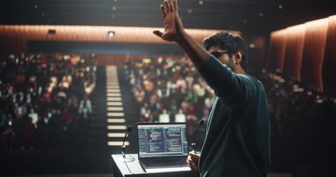 Software Company Lead Developer Making an Inspiring Presentation About Innovation at Technology Conference. Young Indian Man Using Laptop Computer and Talking to an Audience in a Dark Hall
