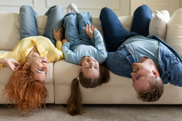 Upside down portrait of joyful family lying on sofa with heads down, mother, father and daughter...