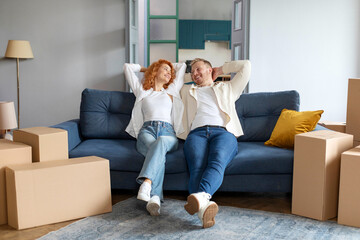 Positive couple having rest on moving day, leaning back on sofa, sitting near carton boxes, looking at each other and smiling, full length shot