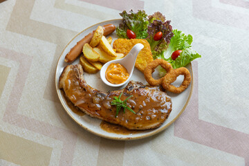 Pork steak, fried onions, sausage, crispy bread with raffles sauce on the steak and dipping sauce in a ceramic plate on a wooden table.