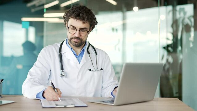 A busy doctor in a white coat writes a prescription using a laptop computer while sitting at a desk at workplace in a modern hospital clinic. Male medical worker physician works, takes notes in office