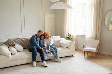 Cheerful parents and kid daughter making selfie on cellphone, having fun and resting on couch at home in large living room, free space