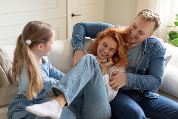 Joyful family of three having fun at home on sofa, man and girl tickling mother and laughing,...