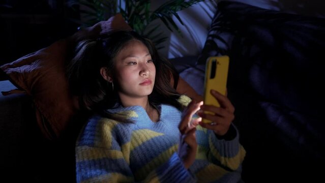 Earnest Asian girl lying on sofa at home using cell phone. Serious discouraged young Chinese woman watching social media app bored on mobile at night. Leisure and online technology in z generation. 