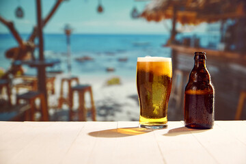 Glass with lager foamy chill beer and beer bottles standing on wooden table at local bar on beach....