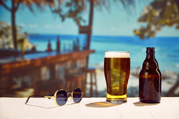 Beachside chills. Frosty beer glass, bottle and sunglasses wooden table, with ocean background on...
