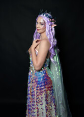 artistic portrait of beautiful female model with long purple hair and elf ears, wearing a fantasy fairy crown, wearing a rainbow glitter sequin ball gown. Standing in side profile, isolated on dark st