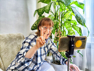 Smiling woman in glasses using a smartphone on a tripod for a video chat. A middle-aged woman...