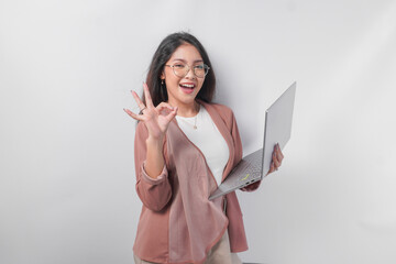 Excited young Asian business woman showing okay sign with right hand and holding laptop on isolated white background.