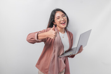 Cheerful young Asian business woman showing thumbs up and holding laptop on isolated white background.