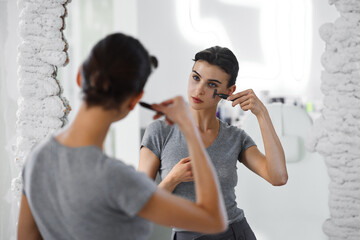 A young woman massages her face with gouache while looking in the mirror.