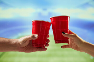 Hands holding red mugs with beer and clicking with blurred sport open air stadium on background....