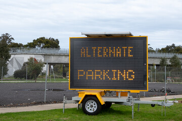 An LED sign mounted on a trailer by the roadside displays the text "alternate parking" outside a closed outdoor car park in an urban area.
