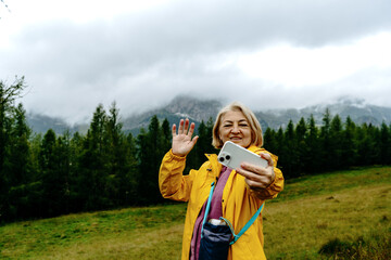 A mature woman in a yellow rain jacket smiles and waves while taking a selfie in a foggy, mountainous forest.