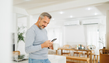 Smiling mature man with grey hair enjoying a cup of coffee and smartphone in a cozy kitchen setting