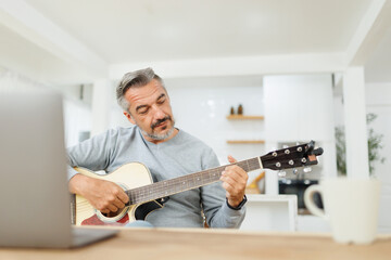 Senior man practicing guitar chords, engaged and thoughtful in a stylishly decorated kitchen with laptop.