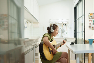 Happy Asian Woman Playing Acoustic Guitar and Singing Along to Music from digital Tablet in a...
