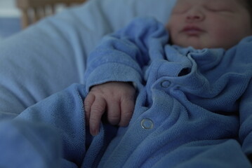 A close-up of a newborn baby’s hand resting on a blue onesie. The tiny fingers and delicate skin...