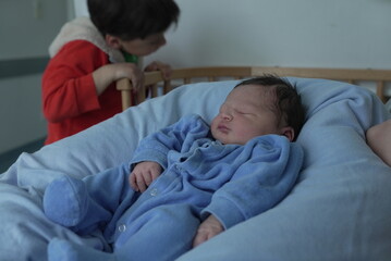 A newborn baby sleeps soundly in a blue onesie on a soft blue blanket while an older child in a red...