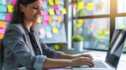 Photograph of a smart, smiling businesswoman working on a laptop computer with many post-it notes on the whiteboard in the background